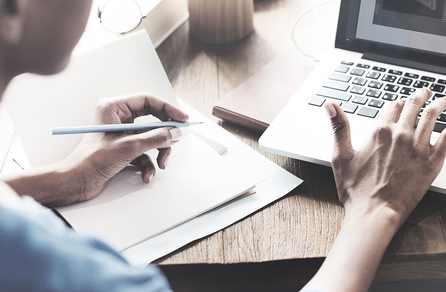 Pessoa digitando no notebook com a mão direita e escrevendo no papel com a mão esquerda