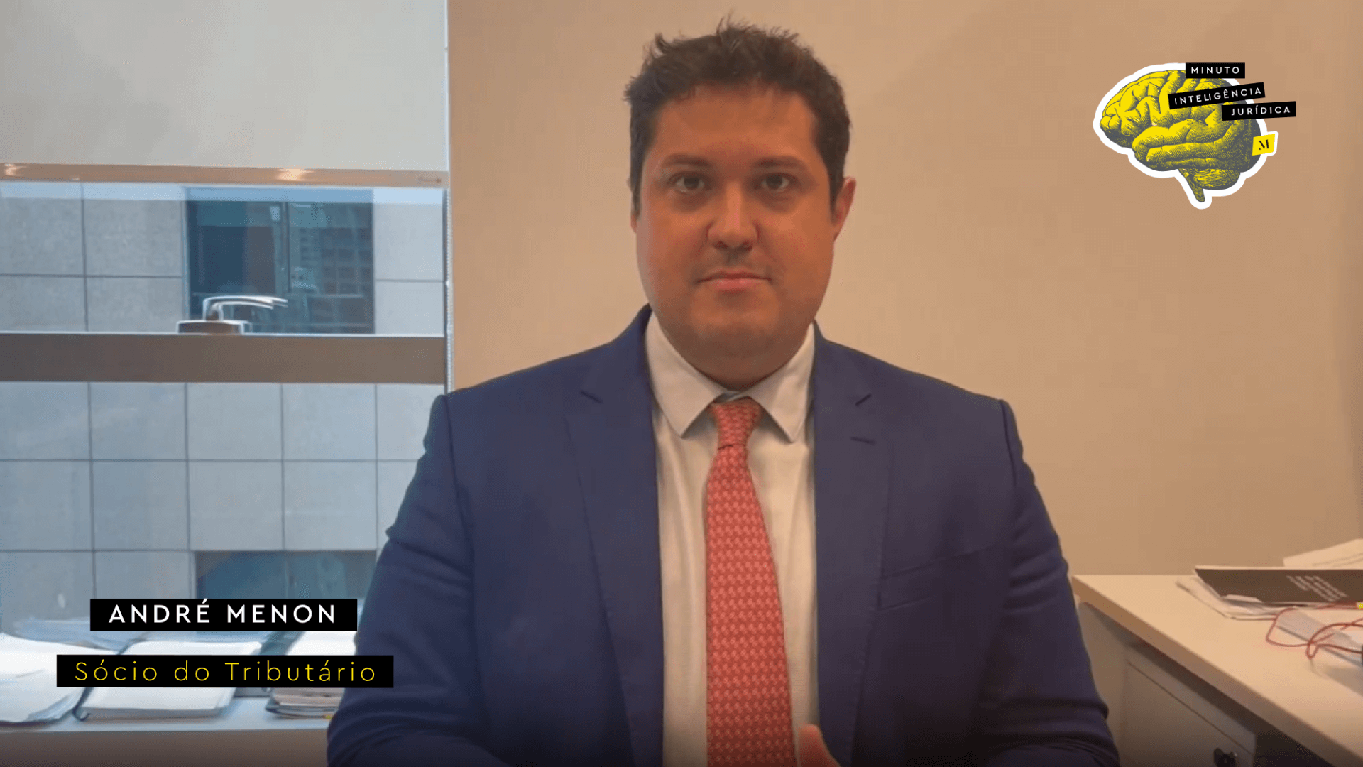 Homem com cabelo preto, usando camisa branca, gravata salmão com bolinhas brancas e blazer azul marinho. No canto inferior esquerdo, faixa preta com frase nas cores branco e amarelo escrito: André Menon, sóciao do Tributário. No canto superior direito, imagem ilustrativa de cérebro amarelo com faixa de texto escrito: Minuto Inteligência Jurídica
