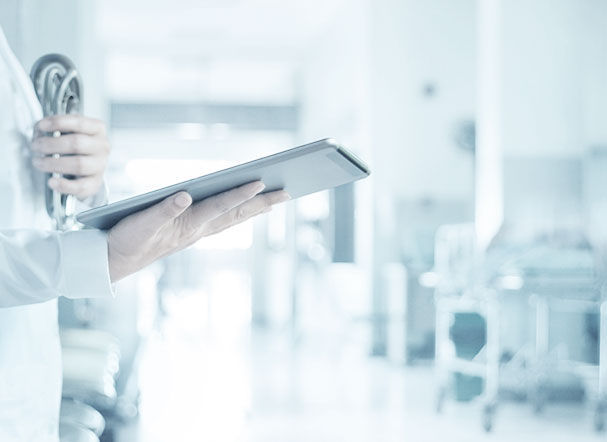 Médica usando jaleco branco, segurando um tablet em uma mão e um estetoscópio na outra.