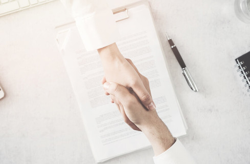 Two white people holding hands as a form of "agreement". In the background, a contract signed by the parties on a table
