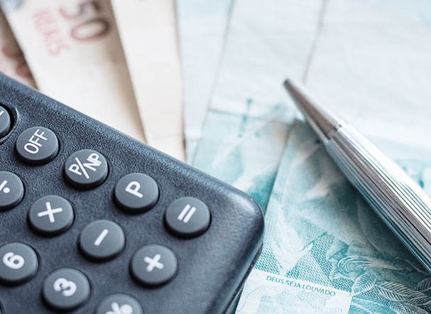 calculadora preta posicionada acima de notas de 50 e 100 reais. Do lado direito da calculadora, uma caneta prata.